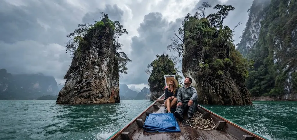 Two passengers in a long-tail boat near karst pillars under a cloudy sky
