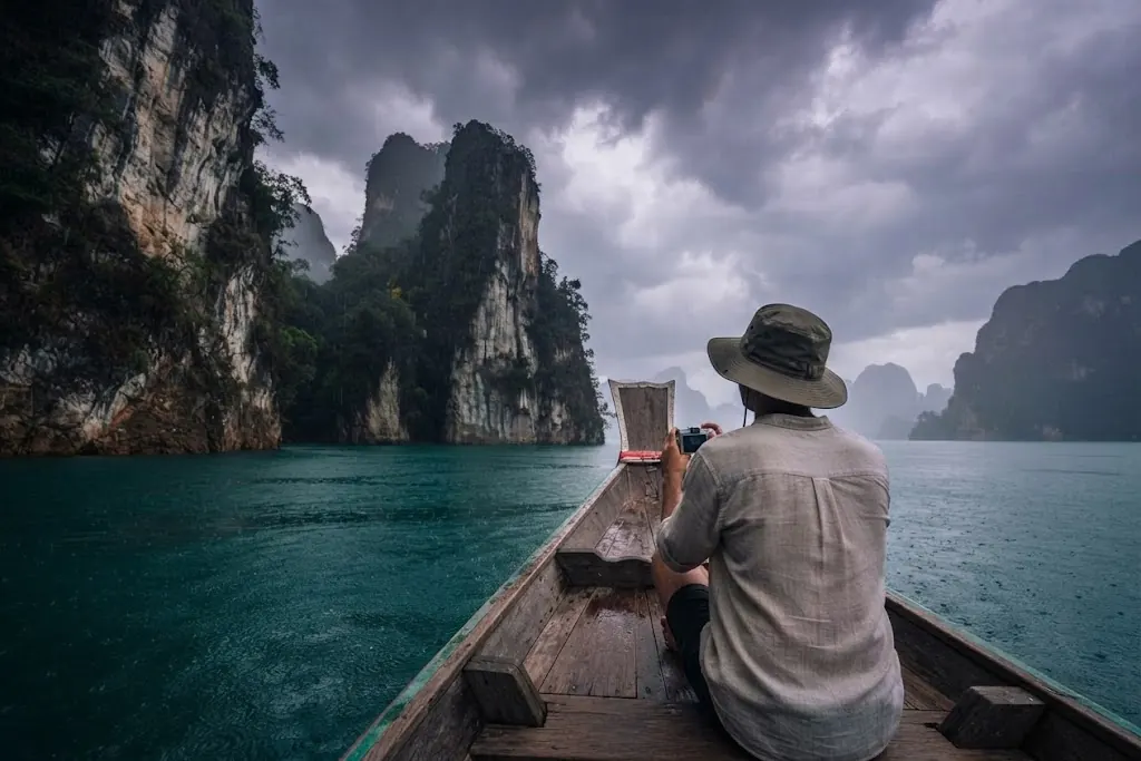 Traveller in a bucket hat photographing karst pillars from a long-tail boat under dark clouds